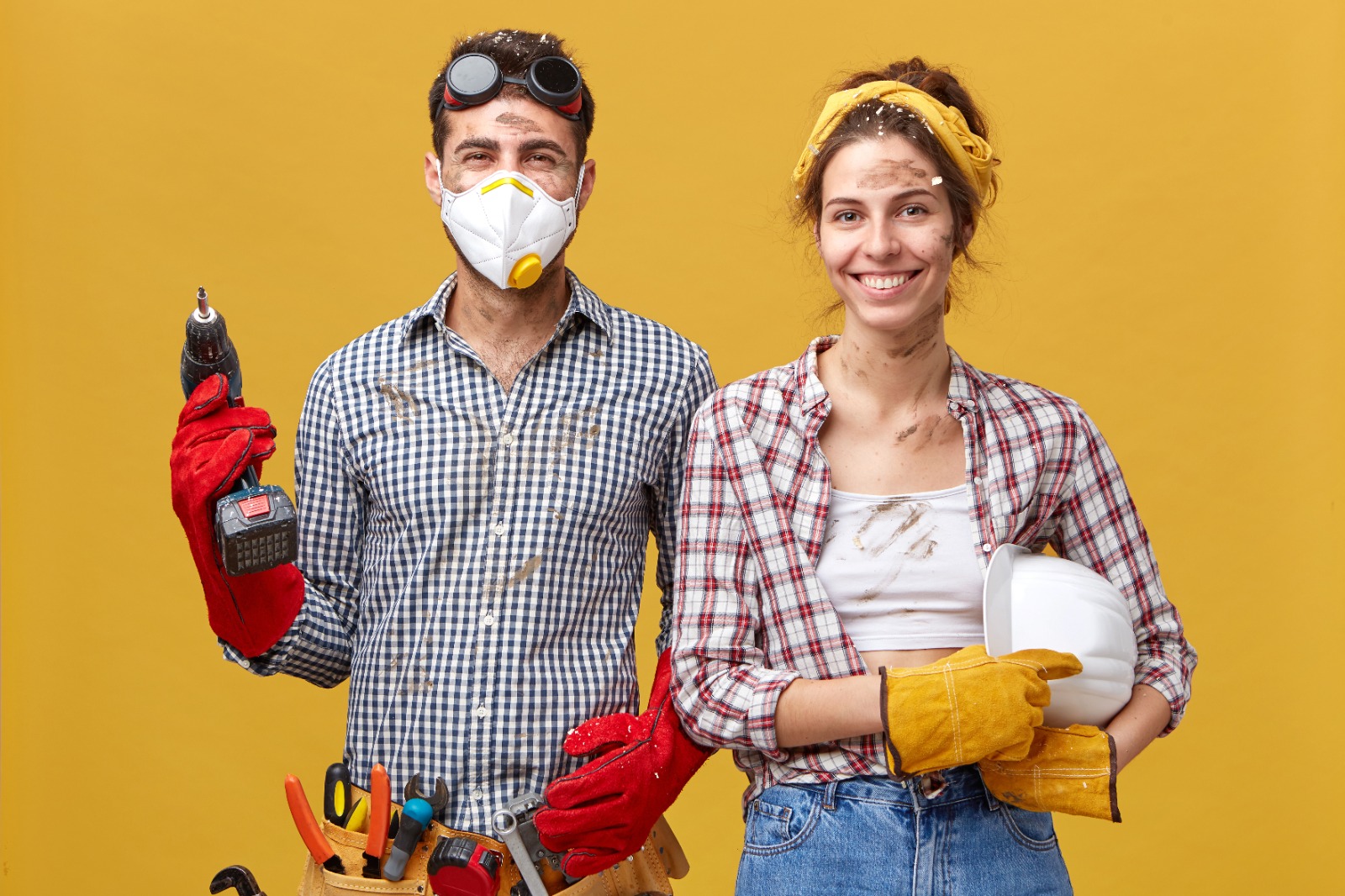 Two tradespeople in work gear, one holding a cordless drill and the other a hard hat, smiling after a job.
